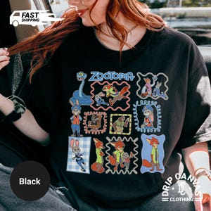 May include: Black t-shirt featuring a colorful graphic of characters from the animated movie "Zootopia." The design includes various characters in framed illustrations, with the movie title at the top. The shirt has a crew neck.