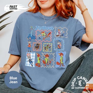 May include: Blue jean colored t-shirt featuring multiple cartoon character illustrations from the movie "Zootopia". The shirt has the word "Zootopia" in a blue and red font. The shirt is made of a soft material.