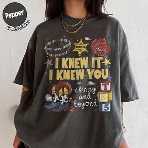 以下が含まれることがあります： I KNEW IT I KNEW YOU infinity and beyondというテキストがプリントされたチャコールグレーのTシャツ。デザインには、カラフルなビーズのブレスレット、黄色の保安官バッジ、赤いカウボーイハット、牛柄のブーツ、3つの積み木が描かれています。