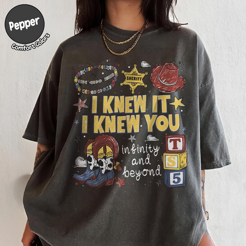 以下が含まれることがあります： I KNEW IT I KNEW YOU infinity and beyondというテキストがプリントされたチャコールグレーのTシャツ。デザインには、カラフルなビーズのブレスレット、黄色の保安官バッジ、赤いカウボーイハット、牛柄のブーツ、3つの積み木が描かれています。