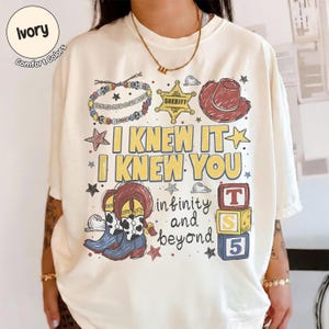 以下が含まれることがあります： アイボリーのTシャツで、I KNEW IT I KNEW YOUおよびinfinity and beyondというテキストのグラフィックプリントが施されています。デザインには、ビーズのブレスレット、保安官の星、赤いカウボーイハット、カウボーイブーツ、3つの積み重ねられたアルファベットブロックが含まれています。