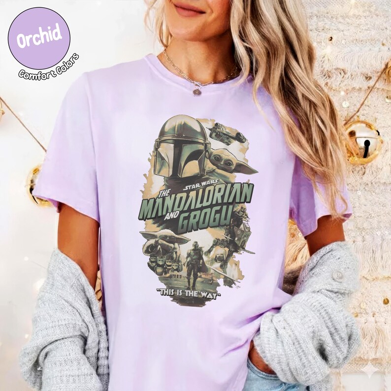 Pu&ograve; includere: Una maglietta lilla con una grafica della serie Star Wars, "The Mandalorian and Grogu". Il design include immagini di The Mandalorian, Grogu e altri personaggi, con la frase "This is the Way" in basso. La maglietta ha il testo "Orchid Comfort Colors" in un cerchio.
