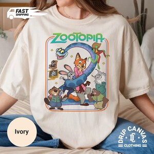 May include: Ivory-colored t-shirt featuring a colorful graphic from the animated movie "Zootopia." The design includes the movie title and various characters like a fox, rabbit, and sloth. The shirt also has the text "Drip Canvas Clothing."