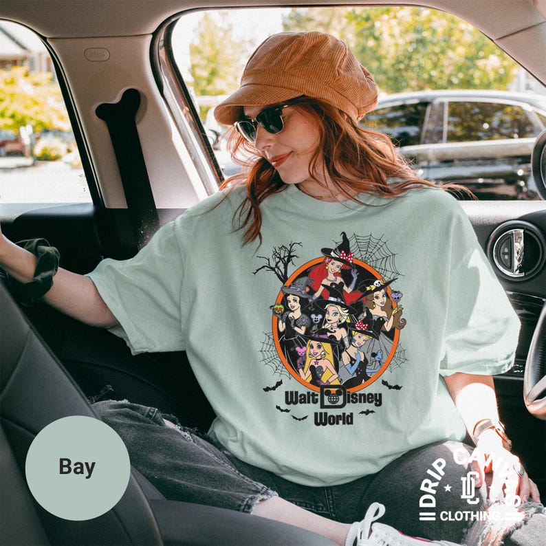 Puede incluir: Camiseta verde claro con un dise&ntilde;o de Halloween de princesas de Disney con sombreros de bruja. El gr&aacute;fico est&aacute; en un c&iacute;rculo naranja con una telara&ntilde;a y las palabras "Walt Disney World".