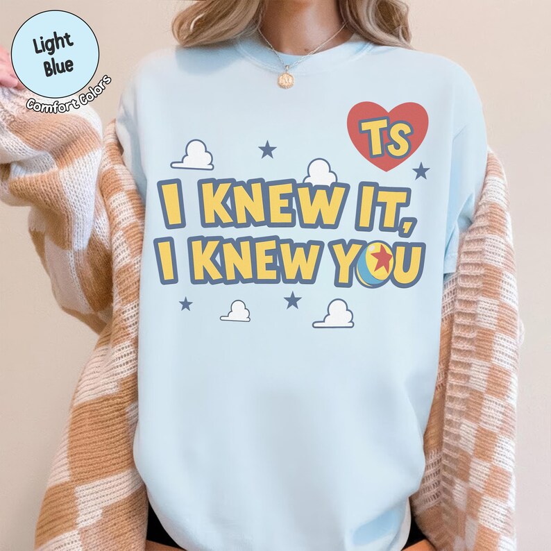 May include: A light blue crewneck sweatshirt featuring a graphic print with the text "I KNEW IT, I KNEW YOU" in yellow letters with a blue outline. The design includes a red heart with "TS" inside, small blue stars, and white cloud shapes.