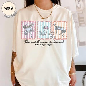 May include: Ivory t-shirt with three framed illustrations of Frozen characters. The illustrations include a bow, a lizard, and a headband with a bow. The text "The cold never bothered me anyway" is below the illustrations.
