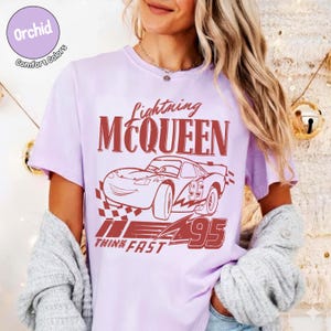 K&ouml;nnte beinhalten: Ein helllila T-Shirt mit einem roten Aufdruck von Lightning McQueen aus dem Film Cars. Der Aufdruck enth&auml;lt den Text "Lightning McQueen", "95" und "Think Fast". Das Shirt ist von der Marke Comfort Colors.
