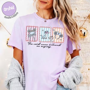 May include: A lilac t-shirt featuring three cartoon images and the text "The cold never bothered me anyway." The images include a bow, a lizard, and mouse ears. The shirt has the words "Orchid Comfort Colors" in a circle.