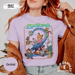 May include: Orchid-colored t-shirt featuring the word "Zootopia" and a graphic of characters from the animated film. The design includes a large number "2" formed by a snake and other characters. The shirt has a crew neck and short sleeves.