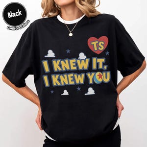 May include: A black short-sleeved t-shirt featuring the yellow text "I KNEW IT, I KNEW YOU" with a red heart containing the letters "TS" and a small ball graphic. The design includes white cloud shapes and small blue stars on a black background.