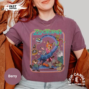 May include: Berry-colored t-shirt featuring a colorful graphic from the movie "Zootopia." The design includes characters like Nick Wilde, Judy Hopps, and others, with the word "Zootopia" in green. The shirt has a crew neck and short sleeves.