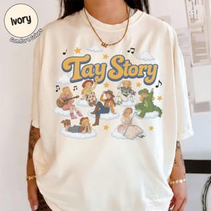 May include: An ivory short-sleeve t-shirt featuring the text Tay Story in a retro yellow font. The graphic includes several illustrated characters dressed in various costumes sitting on white clouds, surrounded by yellow stars and musical notes.