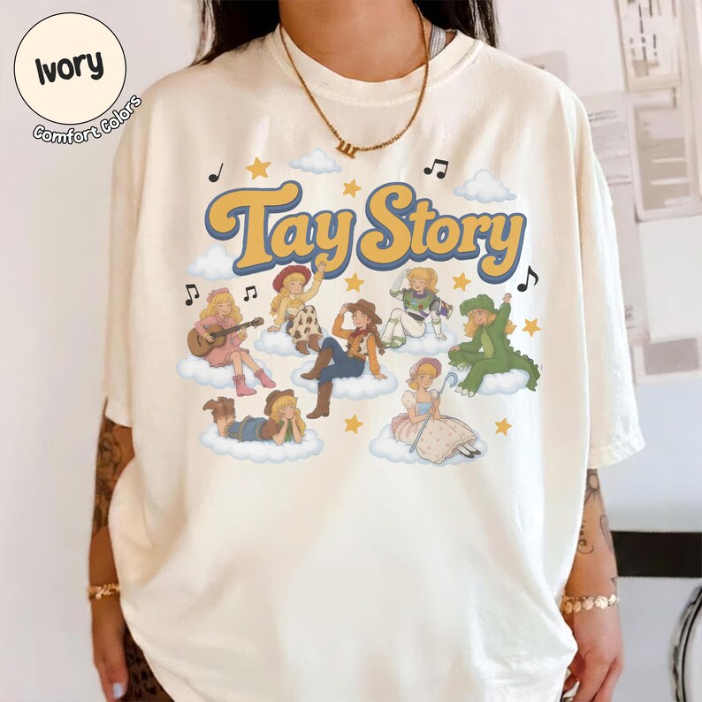May include: An ivory short-sleeve t-shirt featuring the text Tay Story in a retro yellow font. The graphic includes several illustrated characters dressed in various costumes sitting on white clouds, surrounded by yellow stars and musical notes.