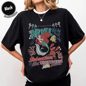May include: A black t-shirt featuring a graphic print of Ariel, Flounder, and Sebastian. The text reads Atlantica Ocean Tour 1991, Ariel, Sebastian and the Hot Crustacean Band. The design includes coral and bubbles in shades of red, teal, and white.