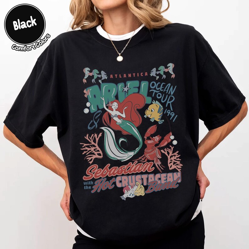 May include: A black t-shirt featuring a graphic print of Ariel, Flounder, and Sebastian. The text reads Atlantica Ocean Tour 1991, Ariel, Sebastian and the Hot Crustacean Band. The design includes coral and bubbles in shades of red, teal, and white.