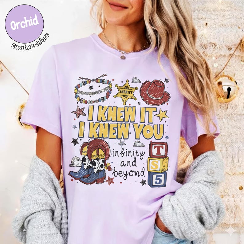 以下が含まれることがあります： 薄紫色のTシャツで、ビーズのブレスレット、黄色い保安官の星、茶色のカウボーイハット、カウボーイブーツのグラフィックプリントが施されています。テキストは「I KNEW IT I KNEW YOU infinity and beyond」と書かれ、T、S、5と書かれた3つの積み木が添えられています。