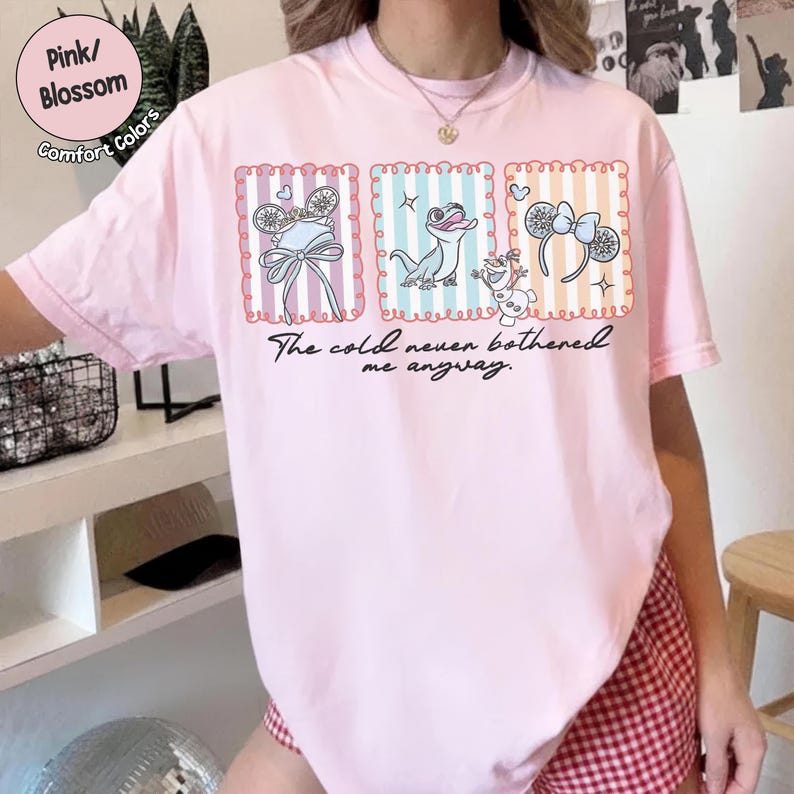 May include: Pale pink t-shirt with a graphic of cartoon characters in frames and the text "The cold never bothered me anyway." The tee has a crew neck and short sleeves. The shirt is labelled "Pink/Blossom Comfort Colors."