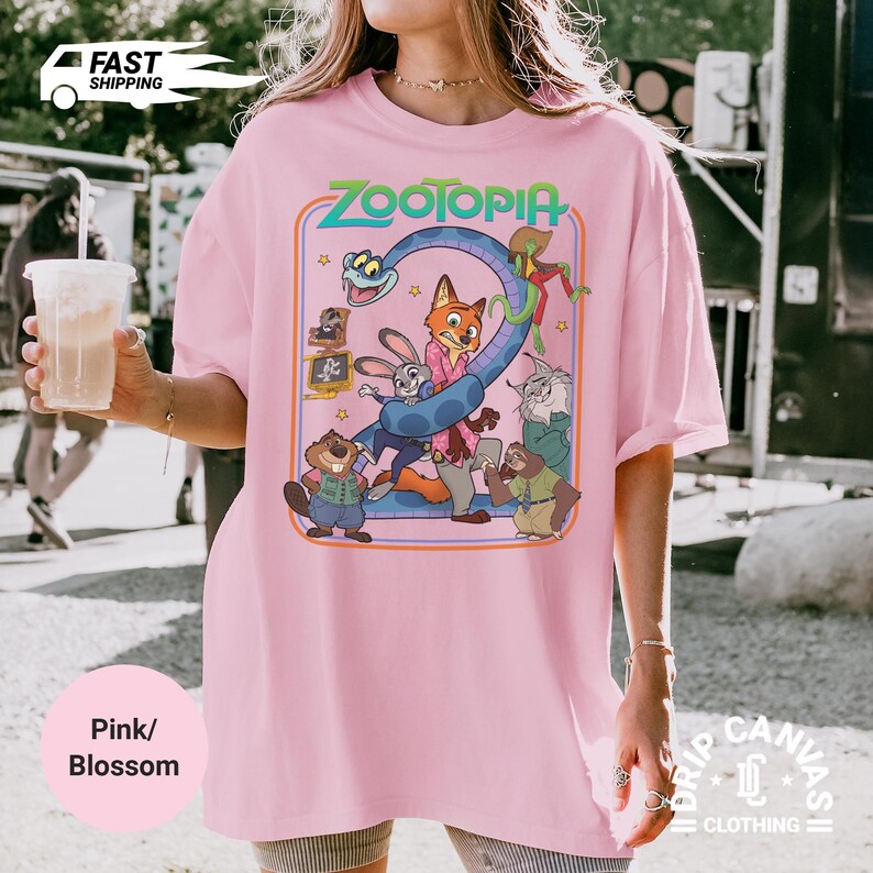 May include: Pink t-shirt with a graphic from the animated movie "Zootopia." The design features the movie's title and several characters, including a fox, a rabbit, and a snake. The shirt has short sleeves and a relaxed fit.