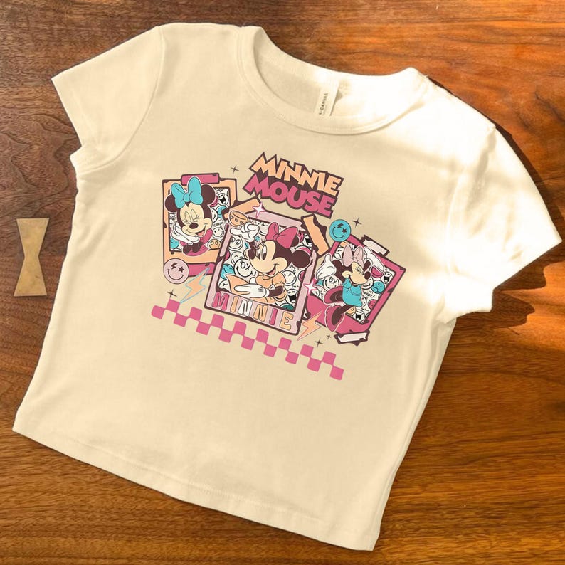May include: Cream-colored t-shirt featuring a graphic of Minnie Mouse in three different poses. The words "MINNIE MOUSE" are printed above the images in a retro font, with a pink and white checkered pattern at the bottom.