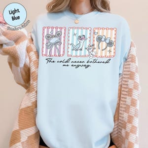 May include: Pale blue Comfort Colors t-shirt with a graphic design. The design features three framed images of a bow, a lizard, and a snowman, with the text "The cold never bothered me anyway."