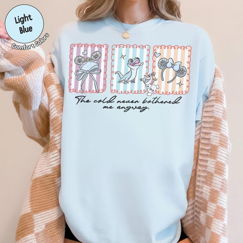 May include: Pale blue Comfort Colors t-shirt with a graphic design. The design features three framed images of a bow, a lizard, and a snowman, with the text "The cold never bothered me anyway."