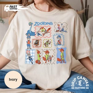 May include: Ivory-colored t-shirt featuring colorful cartoon illustrations of characters from the movie "Zootopia." The shirt includes the movie title and various character portraits within decorative frames. The shirt is part of the "Drip Canvas Clothing" collection.