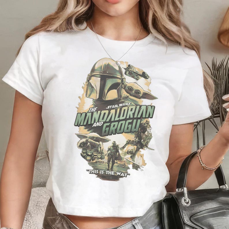Pu&ograve; includere: T-shirt bianca con grafica della serie Star Wars, "The Mandalorian and Grogu". Il design include il casco Mandaloriano, Grogu e la frase "This is the Way". L'opera d'arte &egrave; nei toni del verde, marrone e giallo.