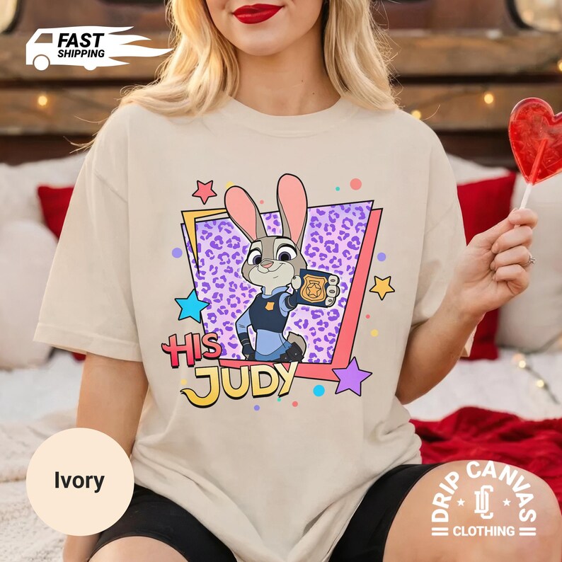May include: Ivory-colored t-shirt featuring a cartoon image of Judy Hopps from Zootopia, holding a badge. The design includes the text "HIS JUDY" in colorful lettering, surrounded by stars and a purple leopard print background. The shirt is from Drip Canvas Clothing.