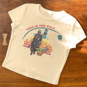 May include: Cream-colored t-shirt featuring a graphic of The Mandalorian and Grogu, with the text "THIS IS THE WAY" above them. The design includes a colorful sunset-style background and a spaceship icon.