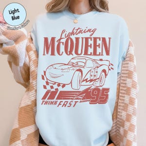 K&ouml;nnte beinhalten: Hellblaues T-Shirt mit roter Grafik des Rennwagens Lightning McQueen und dem Text "Lightning McQueen". Die Nummer "95" befindet sich auf dem Auto und die Worte "Think Fast" stehen darunter.