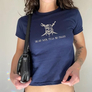 May include: Navy blue t-shirt featuring a skull with crossed swords and the text "DEAD MEN TELL NO TALES" in white. The shirt has short sleeves and a crew neck. A black shoulder bag is visible.