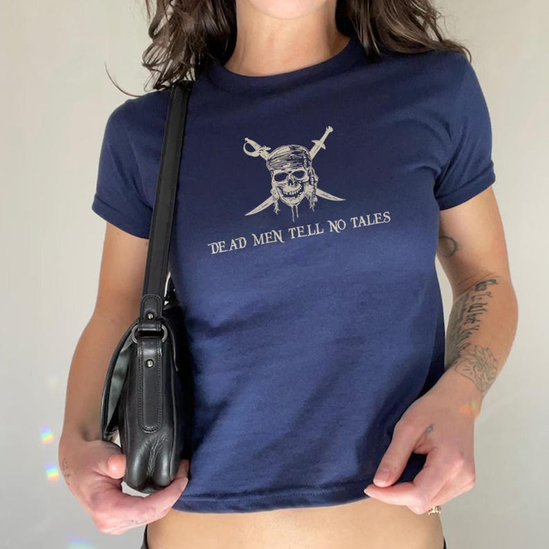 May include: Navy blue t-shirt featuring a skull with crossed swords and the text "DEAD MEN TELL NO TALES" in white. The shirt has short sleeves and a crew neck. A black shoulder bag is visible.