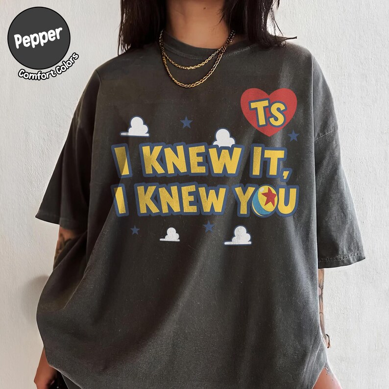 May include: A charcoal gray short-sleeve t-shirt featuring the text "I KNEW IT, I KNEW YOU" in yellow with blue outlines. The design includes a red heart with "TS" inside, small blue stars, white clouds, and a ball with a red star graphic.