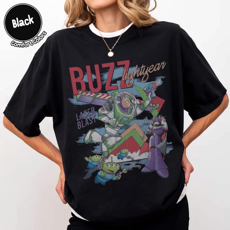 Może przedstawiać: Czarny t-shirt z nadrukiem graficznym przedstawiającym Buzza Astrala, Zurga i trzy zielone ufoludki. Napis głosi "BUZZ lightyear" na czerwono i biało, "LASER BLAST" na niebiesko oraz "Aliens" na biało. Wz&oacute;r zawiera geometryczne kształty w kolorze czerwonym i zielonym.
