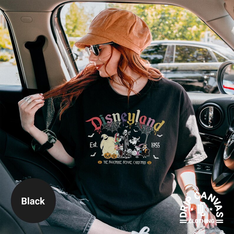 May include: Black t-shirt with a Disneyland graphic featuring characters from The Nightmare Before Christmas. The design includes the text "Disneyland Est. 1955" in a colorful, dripping font, with spiderwebs and bats. The shirt is part of the Drip Canvas Clothing collection.