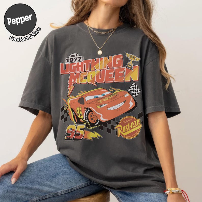 Op de afbeelding: Donkergrijs Lightning McQueen grafisch t-shirt. Het ontwerp toont het personage Lightning McQueen, het nummer 95 en de tekst "Rusteze". Het shirt heeft korte mouwen en een ronde hals.