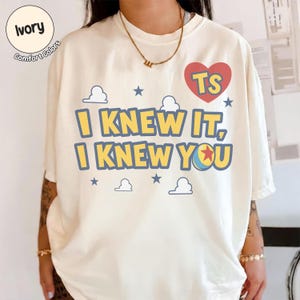 May include: An ivory short-sleeve t-shirt featuring the text I KNEW IT, I KNEW YOU in yellow with blue outlines. The design includes a red heart with the letters TS, small blue stars, white cloud outlines, and a colorful ball with a red star.