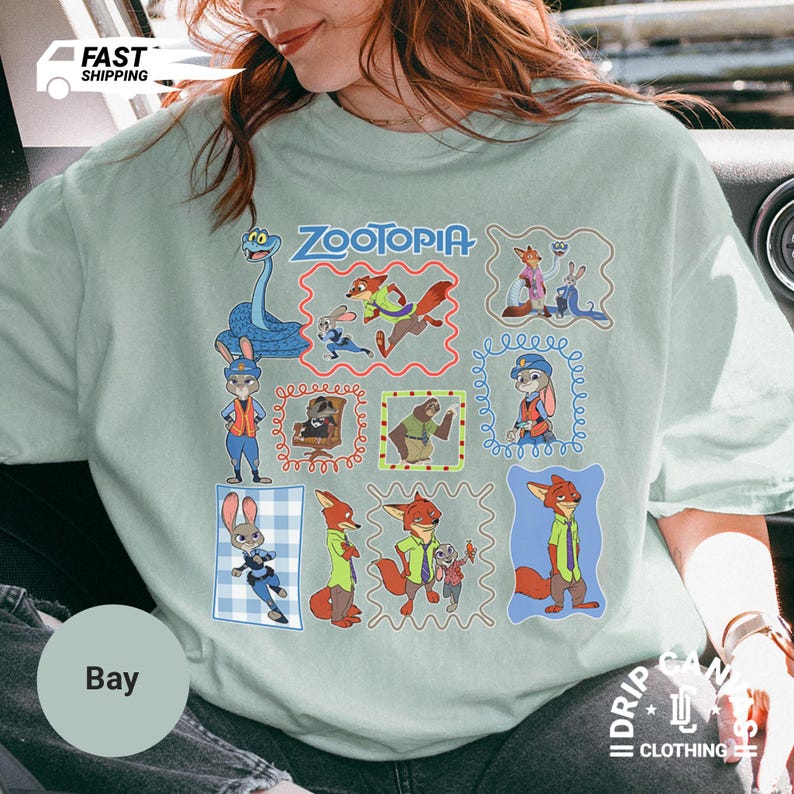 May include: A light green t-shirt featuring the word "ZOOTOPIA" above a grid of cartoon character illustrations. The illustrations include a blue giraffe, a fox, a rabbit, and other characters from the animated movie.