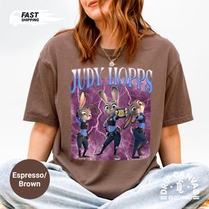 May include: Espresso brown t-shirt featuring a graphic of Judy Hopps from Zootopia. The design includes the text "JUDY HOPPS" in blue and a purple lightning bolt background. The image shows Judy Hopps in a police uniform.