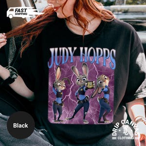 May include: Black t-shirt featuring a graphic of Judy Hopps from Zootopia. The design includes the text "JUDY HOPPS" in blue and white, with three images of Judy Hopps in her police uniform against a purple lightning background.