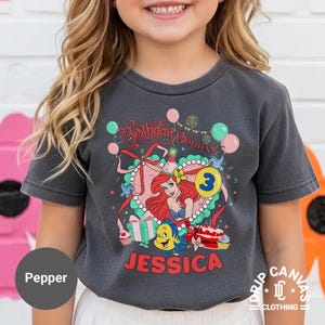 May include: Dark gray t-shirt featuring a colorful graphic of a princess with red hair, a blue tail, and the number 3. The shirt has the text "Birthday Princess" and the name "Jessica". The shirt is from Drip Canvas Clothing.