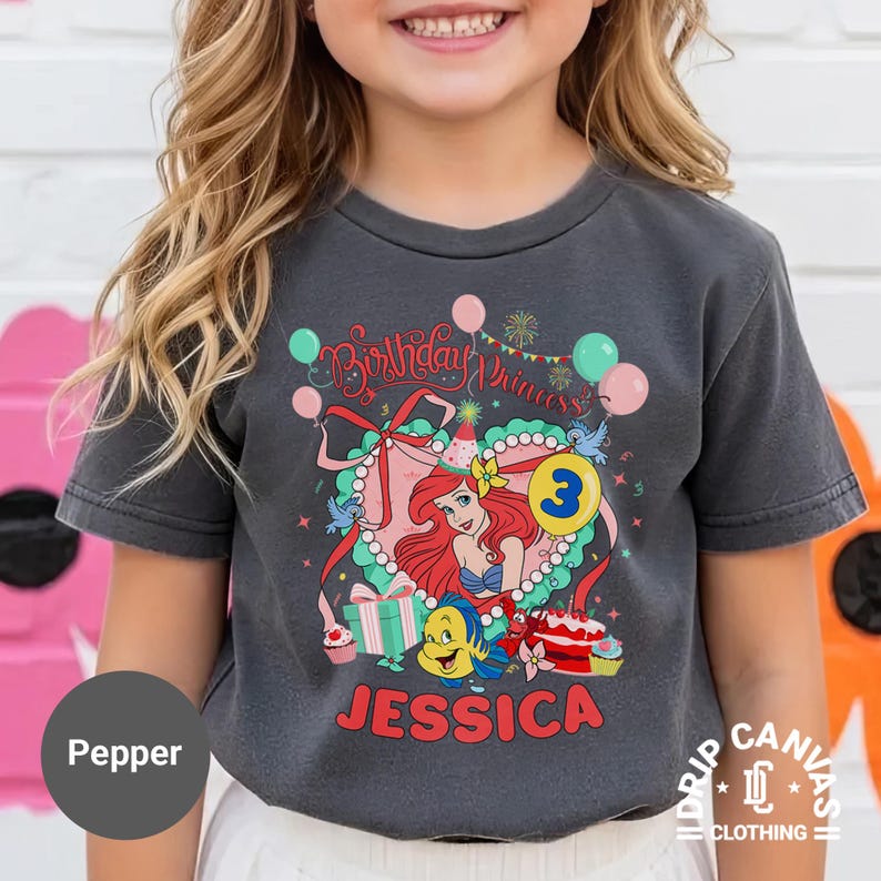 May include: Dark gray t-shirt featuring a colorful graphic of a princess with red hair, a blue tail, and the number 3. The shirt has the text "Birthday Princess" and the name "Jessica". The shirt is from Drip Canvas Clothing.