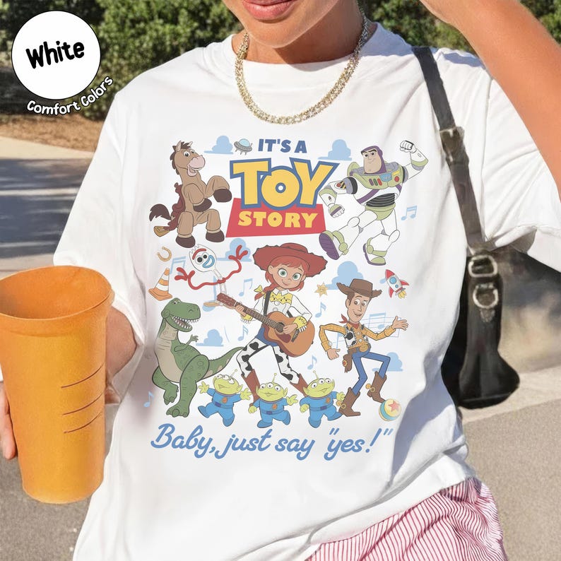Op de afbeelding: Een wit t-shirt met een kleurrijke grafische print van Toy Story-personages, waaronder Woody, Buzz Lightyear, Jessie, Bullseye, Rex, Forky en de Aliens. De tekst luidt IT'S A TOY STORY en Baby, just say "yes!" in blauw en rood.