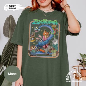May include: Moss green t-shirt featuring a colorful graphic from the animated movie "Zootopia." The design includes characters like Judy Hopps, Nick Wilde, and others. The word "Zootopia" is in a stylized font. The shirt is a relaxed fit.