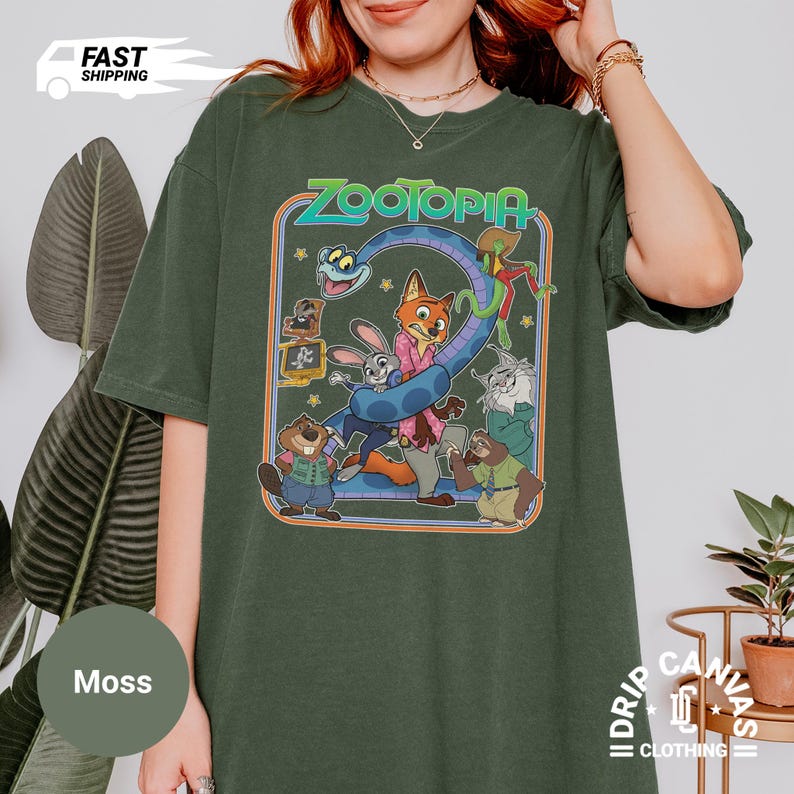 May include: Moss green t-shirt featuring a colorful graphic from the animated movie "Zootopia." The design includes characters like Judy Hopps, Nick Wilde, and others. The word "Zootopia" is in a stylized font. The shirt is a relaxed fit.