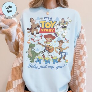 Op de afbeelding: Een lichtblauw sweatshirt met ronde hals en een kleurrijke grafische print van Toy Story-personages, waaronder Woody, Buzz Lightyear, Jessie, Bullseye, Rex, Forky en drie aliens. De tekst luidt IT'S A TOY STORY en Baby, just say yes!