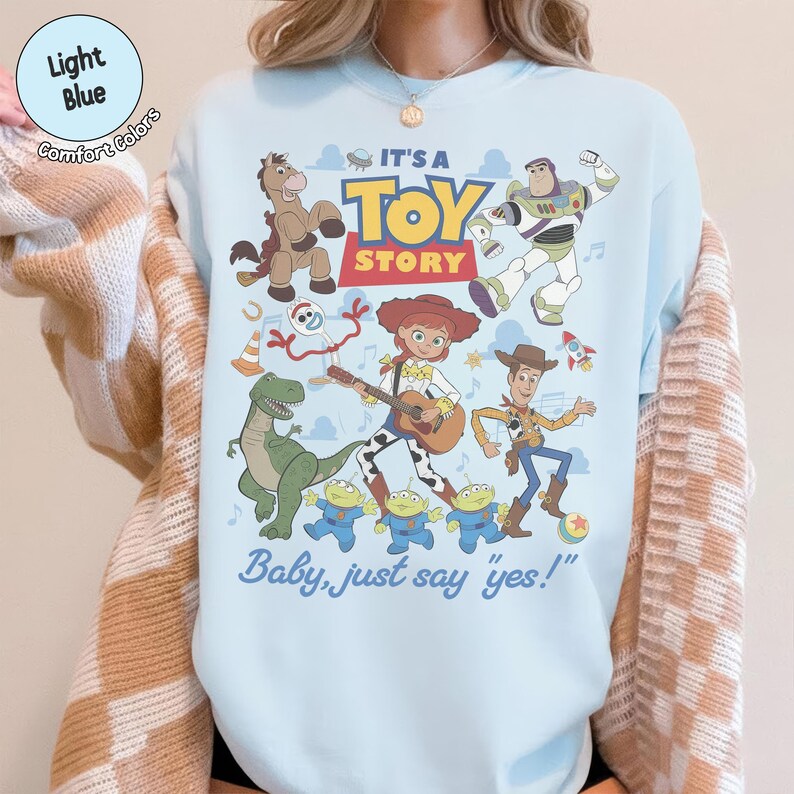 Op de afbeelding: Een lichtblauw sweatshirt met ronde hals en een kleurrijke grafische print van Toy Story-personages, waaronder Woody, Buzz Lightyear, Jessie, Bullseye, Rex, Forky en drie aliens. De tekst luidt IT'S A TOY STORY en Baby, just say yes!