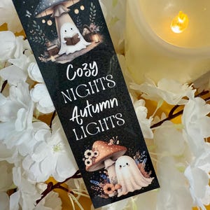 May include: A black bookmark featuring the text "Cozy Nights Autumn Lights" and illustrations of mushrooms and ghosts. The bookmark is decorated with a mushroom and a ghost reading a book. The background is white flowers and a candle.