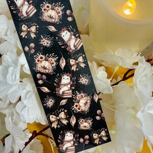 Op de afbeelding: Zwarte washi tape met een herhalend patroon van een spook met een bloemenkroon, pompoenen, strikken en bloemstukken. De tape is versierd met herfstthema-illustraties in tinten bruin, beige en wit.