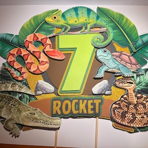 May include: A layered cake topper featuring a large lime green number 7 and the name ROCKET in gold glitter text. The design includes a green chameleon, a brown and orange snake, a blue turtle, a coiled brown snake, and a crocodile among green leaves.